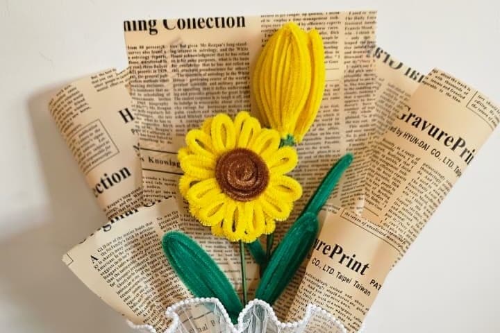 A hand-twisted sunflower with a coiled brown centre, wrapped in newsprint paper with a lace trim