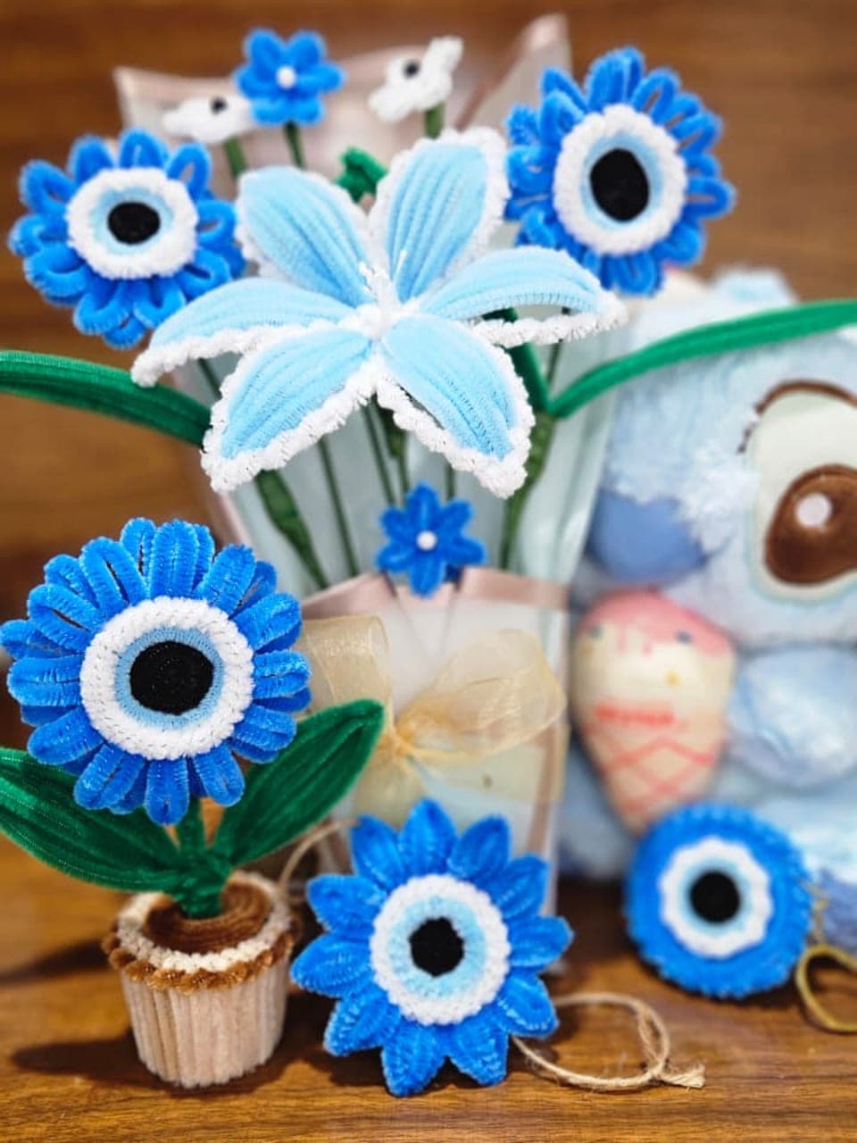 A spread of handcrafted pipe cleaner pieces — evil eye blooms, a lily bouquet, a potted bloom and a keychain, on a wooden table